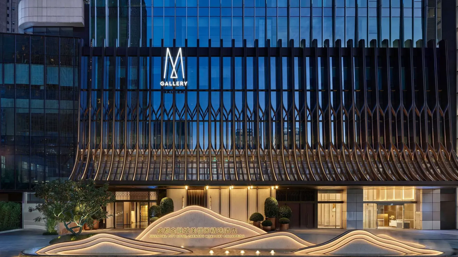 La façade du Financial City Hotel Chengdu – MGallery Collection à la nuit tombée, avec le logo MGallery, son auvent de lames de bronze et la sculpture lumineuse portant le nom de l'hôtel