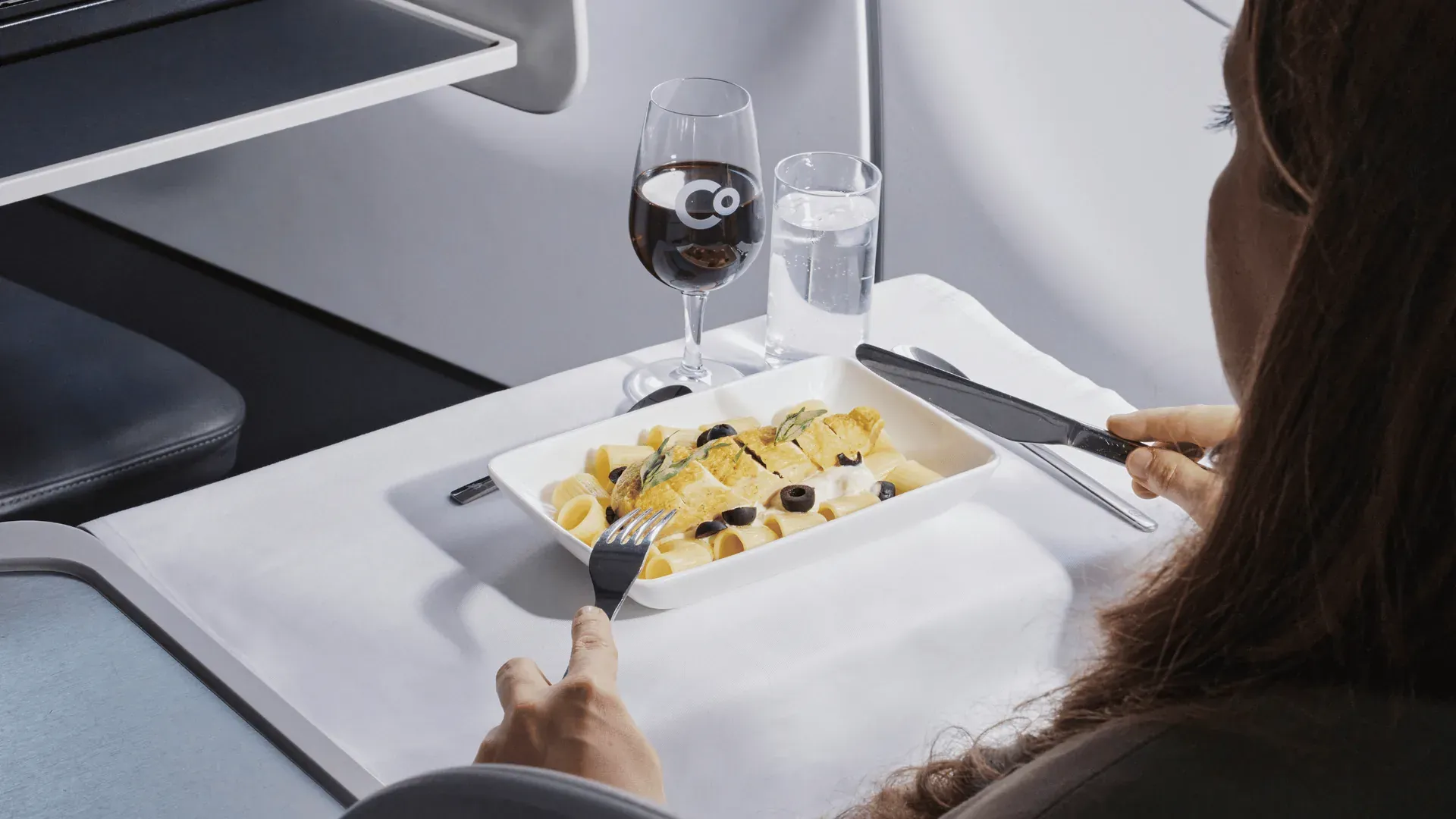Une passagère de La Compagnie attablée à son siège, devant une assiette de pâtes, un verre de vin siglé Co et un verre d'eau posés sur la tablette