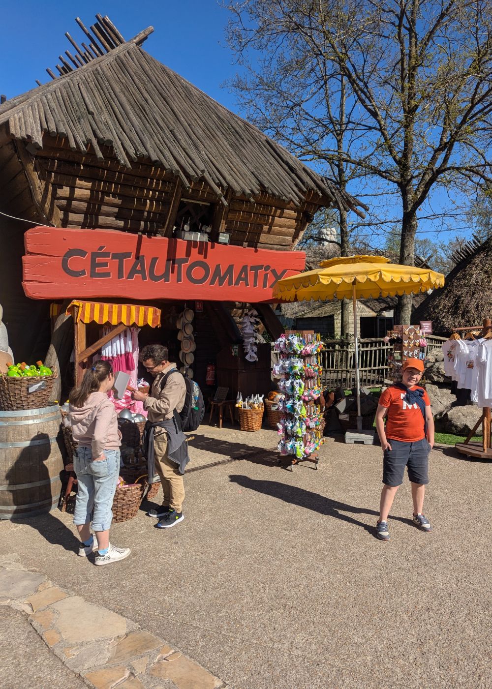 Boutique en bois «&nbsp;Cétautomatix&nbsp;» avec quelques visiteurs observant des souvenirs sous un ciel ensoleillé.