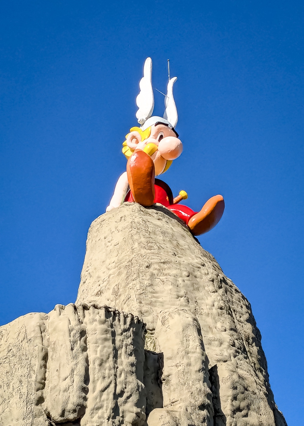 Grande statue d'Astérix assis au sommet d'un rocher beige sous un ciel bleu clair.