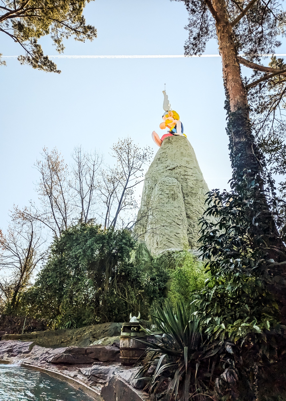Grande statue d'Astérix assis au sommet d'un rocher entouré d'arbres et de végétation.