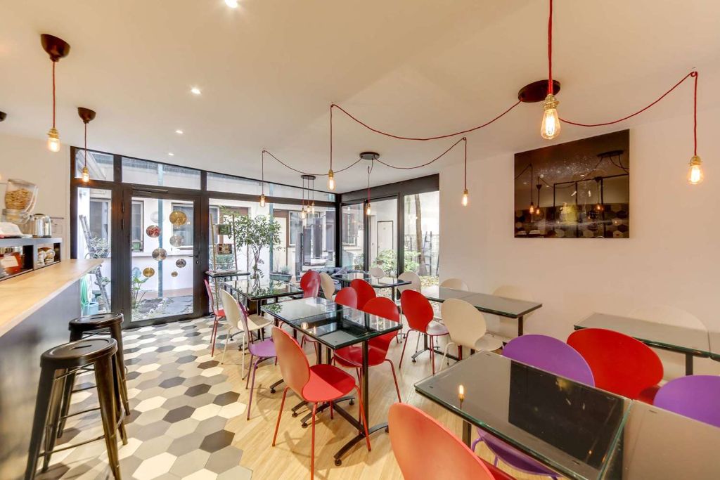 Salle lumineuse avec tables en verre, chaises colorées et ampoules suspendues dans un hôtel parisien.