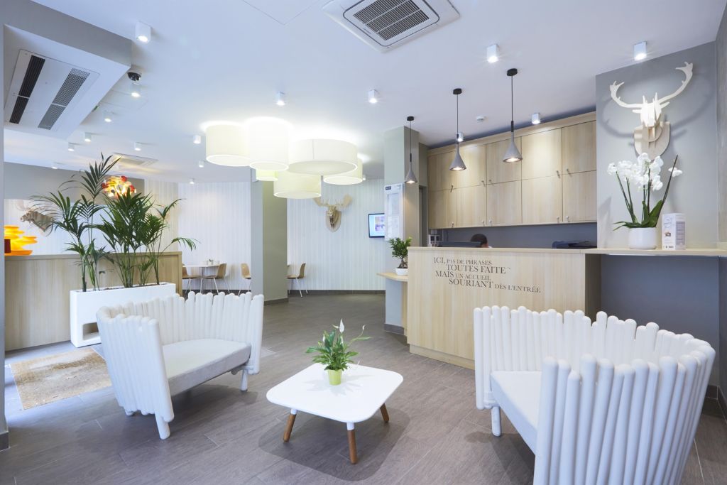 Hall d'accueil lumineux avec fauteuils blancs, comptoir en bois clair et plantes vertes décoratives.