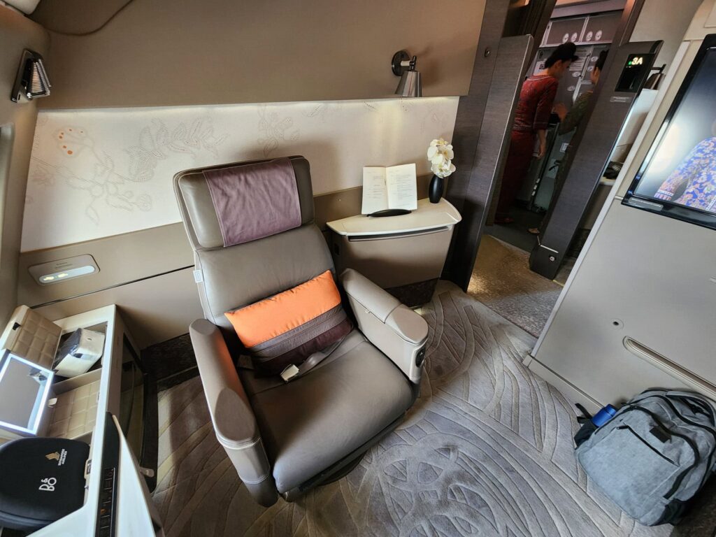Suite Singapore Airlines First Class avec siège inclinable, table et hôtesse en uniforme à l'arrière.