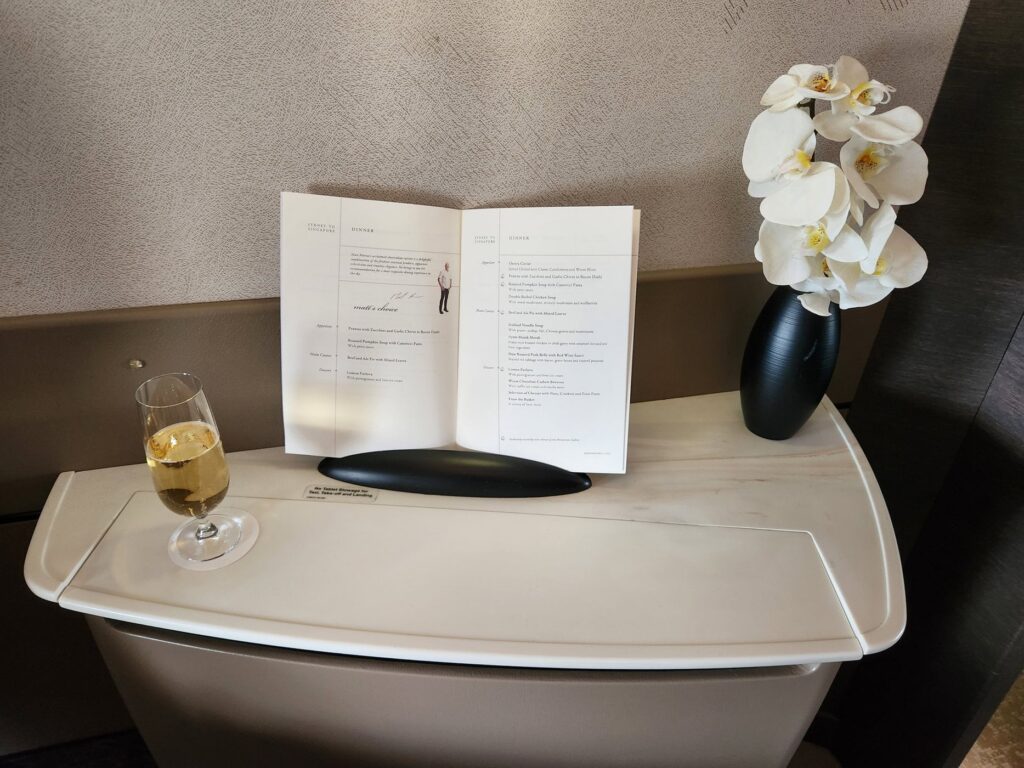 Table d'une suite Singapore Airlines Première Classe avec menu, champagne et orchidée blanche en vase noir.