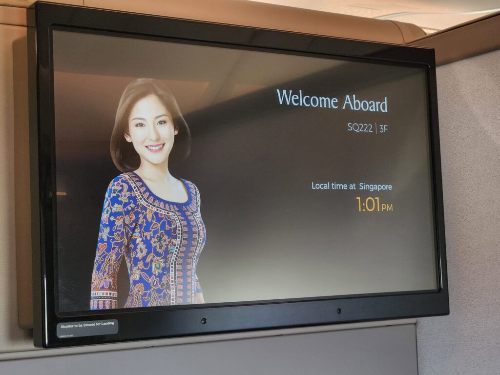 Écran de bienvenue du vol Singapore Airlines SQ222 affichant une hôtesse et l'heure locale à Singapour.
