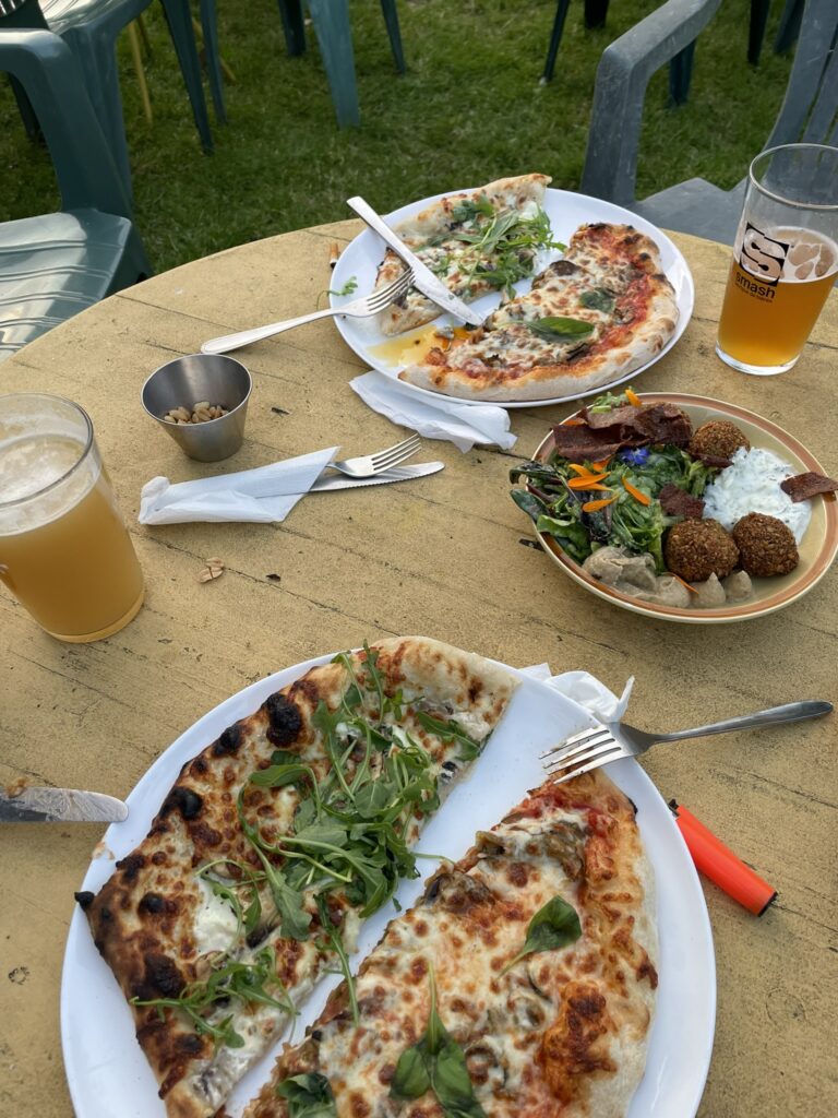 Deux assiettes de pizza garnies de roquette posées sur une table extérieure avec bières artisanales.