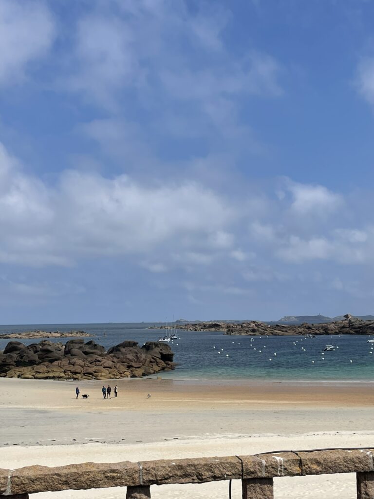 Plage de sable clair avec rochers de granit rose, mer turquoise et quelques promeneurs à marée basse.
