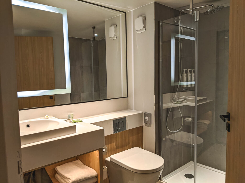 Salle de bain moderne avec grande douche vitrée, miroir éclairé, lavabo carré et toilettes blanches.