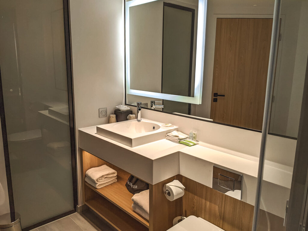 Salle de bain moderne avec grand miroir lumineux, vasque blanche, serviettes pliées et porte en bois clair.