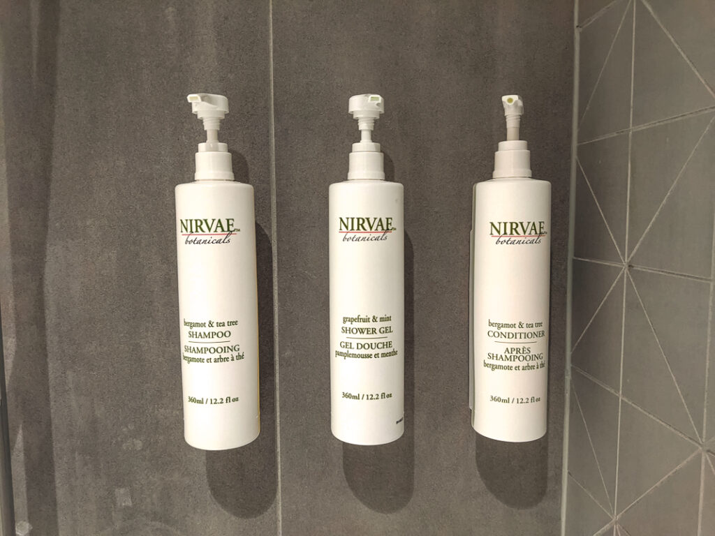 Trois distributeurs muraux Nirvae Botanicals contenant shampoing, gel douche et après-shampoing dans une douche d'hôtel.