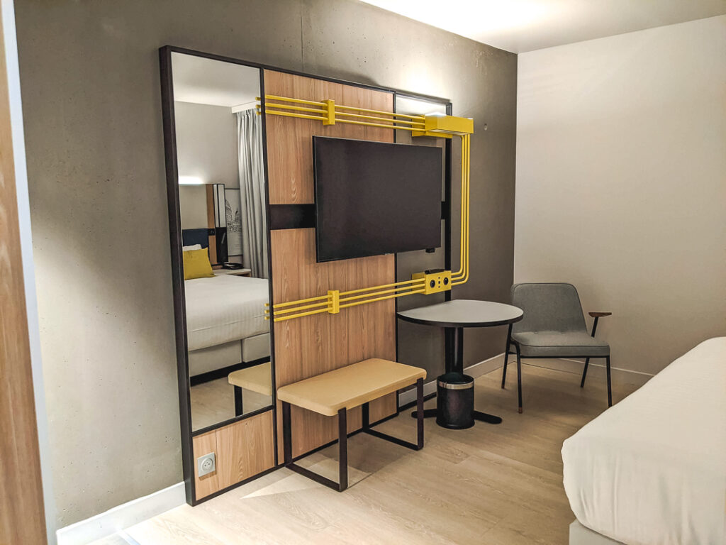 Chambre moderne avec télévision murale, miroir pleine hauteur, chaise grise et accents jaunes au Courtyard Paris Créteil.