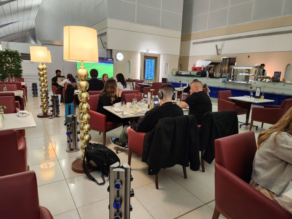 Passagers installés dans le salon Emirates avec sièges rouges et coin restauration avant leur vol.