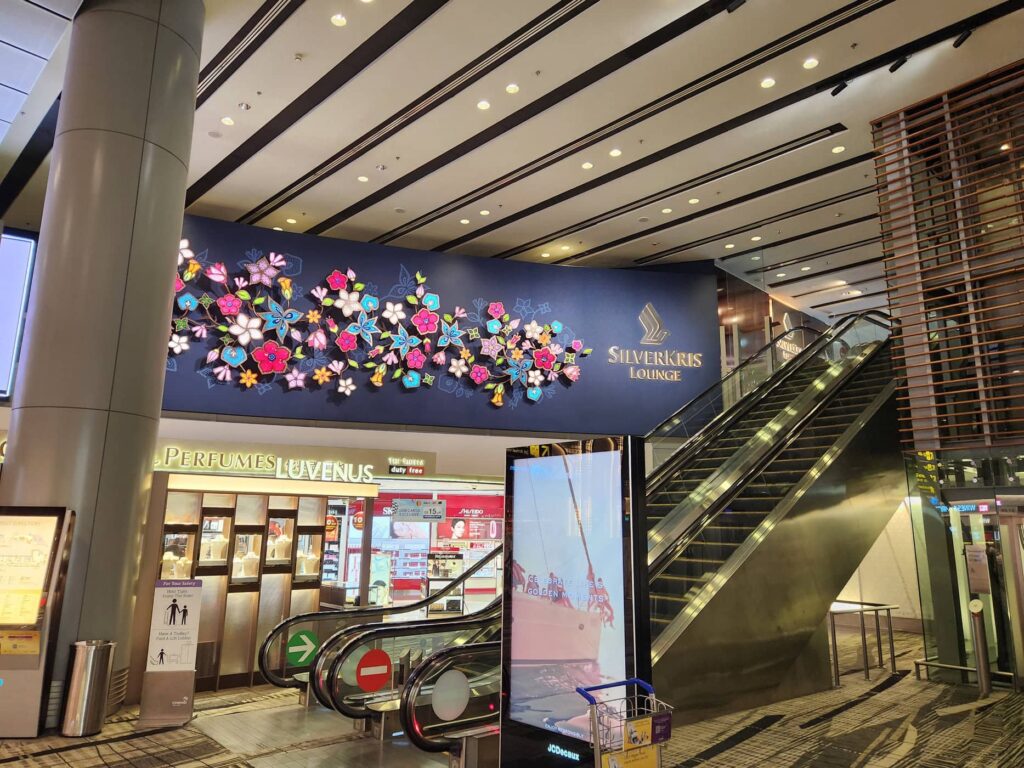 Entrée du salon SilverKris Lounge de Singapore Airlines à l'aéroport avec escalator et décor floral lumineux.