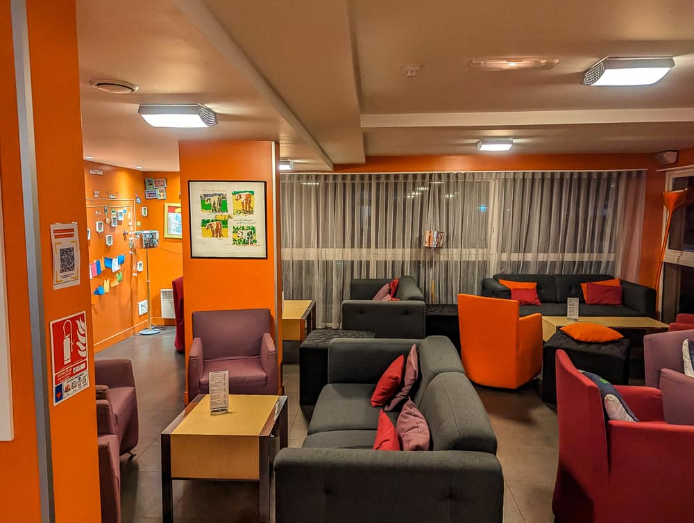 Salon commun coloré avec canapés gris et fauteuils orange à l'hôtel Ibis Styles Ouistreham