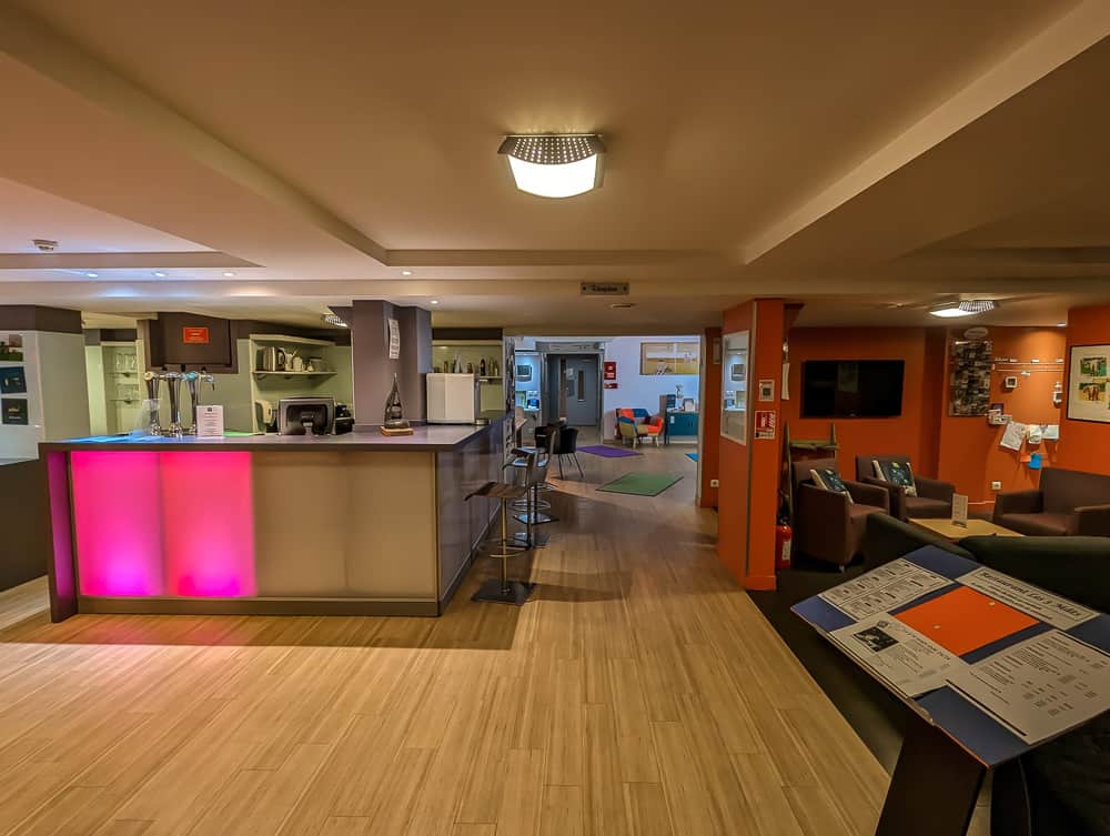 Réception moderne de l'hôtel Ibis Styles Ouistreham avec comptoir éclairé rose et coin salon orangé convivial