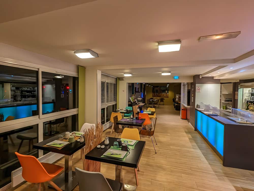 Salle de petit-déjeuner lumineuse avec chaises colorées, tables dressées et comptoir bleu éclairé à l'Ibis Styles