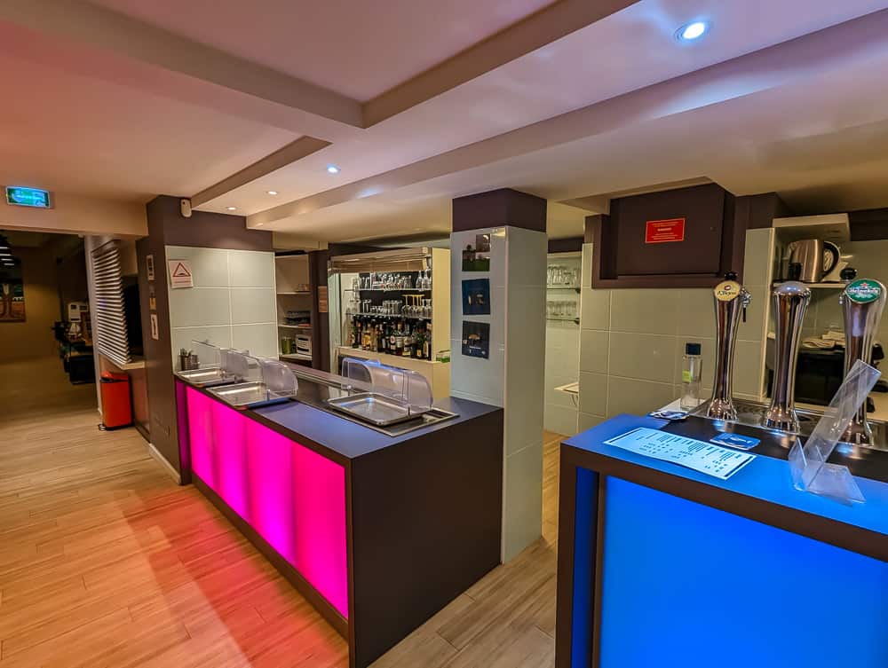 Comptoir de bar moderne avec éclairage rose et bleu, tireuses à bière et étagères de bouteilles à l'Ibis Styles