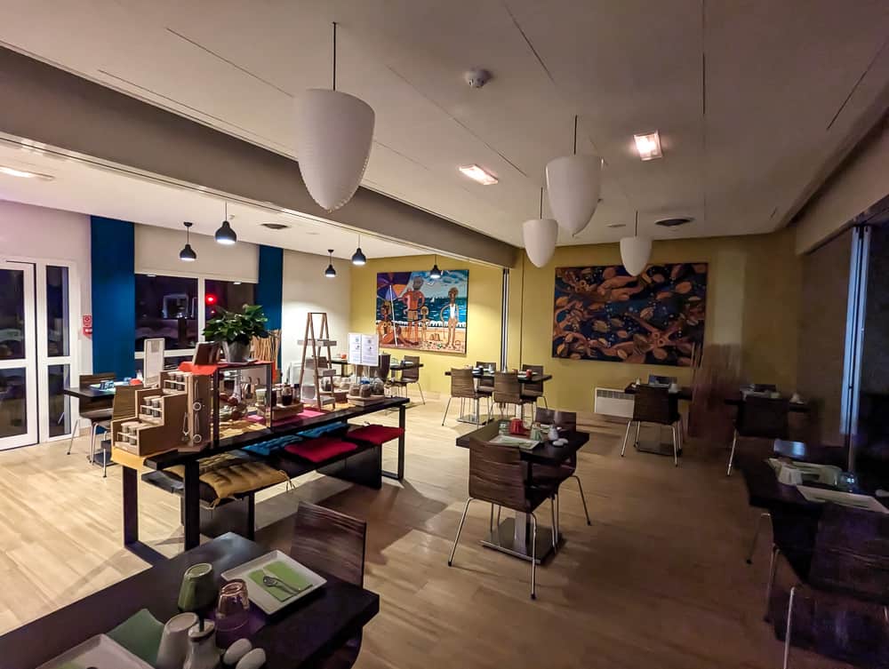 Salle de petit-déjeuner moderne avec tables dressées, buffet central en bois et grandes toiles colorées au mur