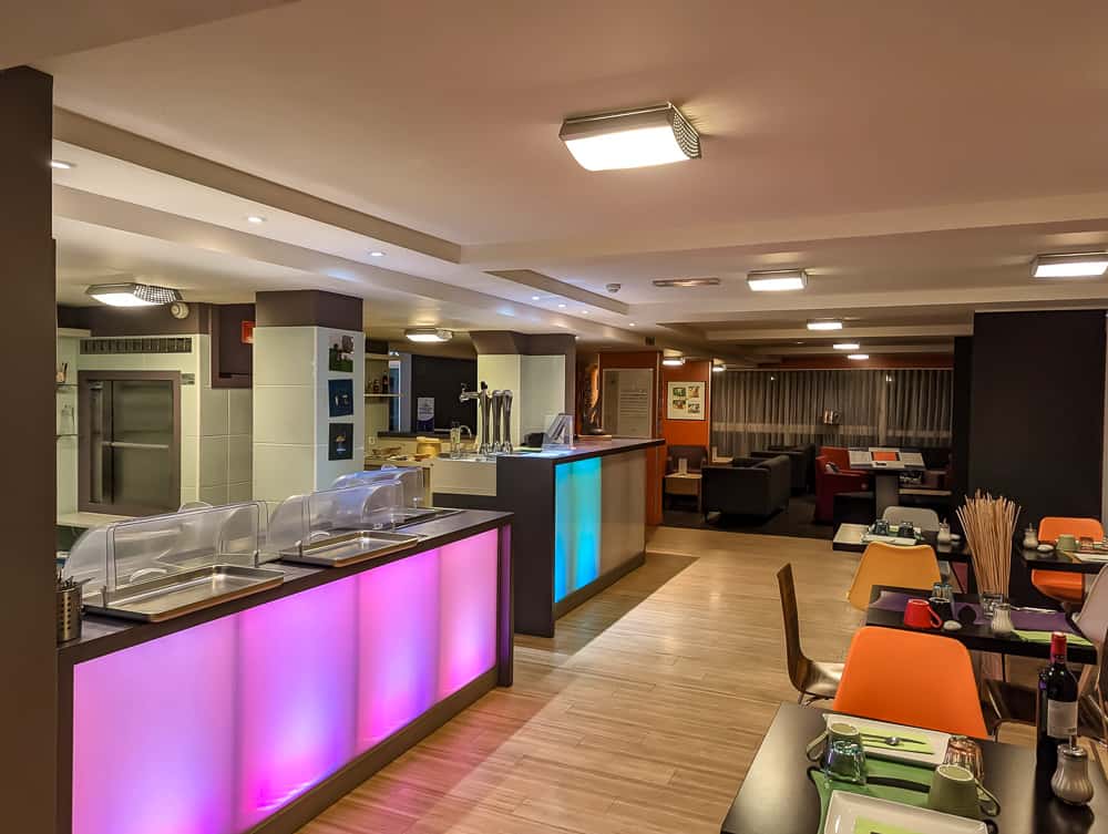 Salle de restaurant moderne avec comptoirs lumineux multicolores et tables dressées à l'Ibis Styles Ouistreham