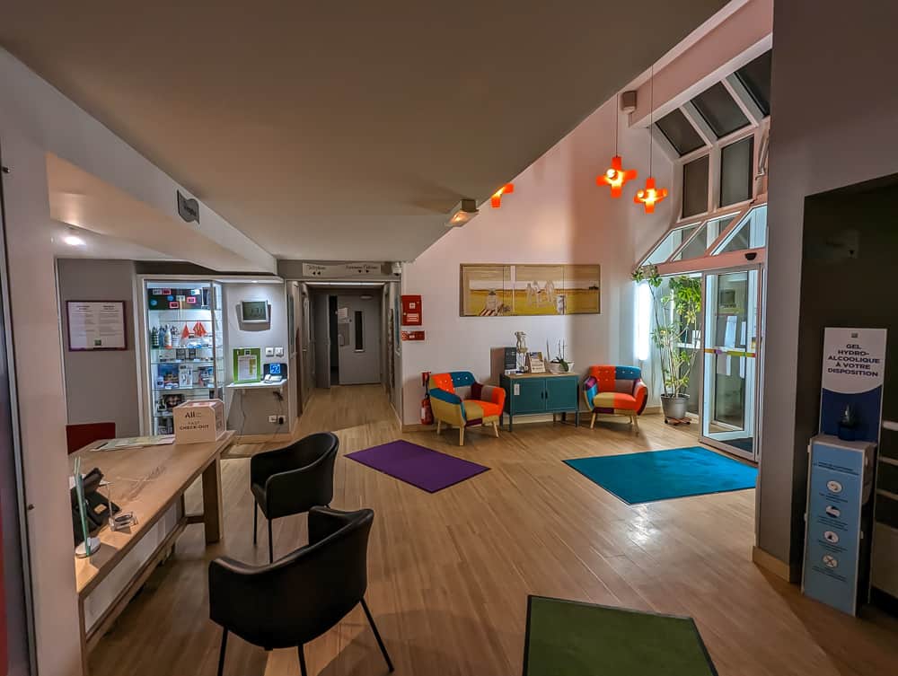 Hall d'accueil lumineux de l'Ibis Styles Ouistreham avec fauteuils colorés, comptoir d'accueil et suspensions orange