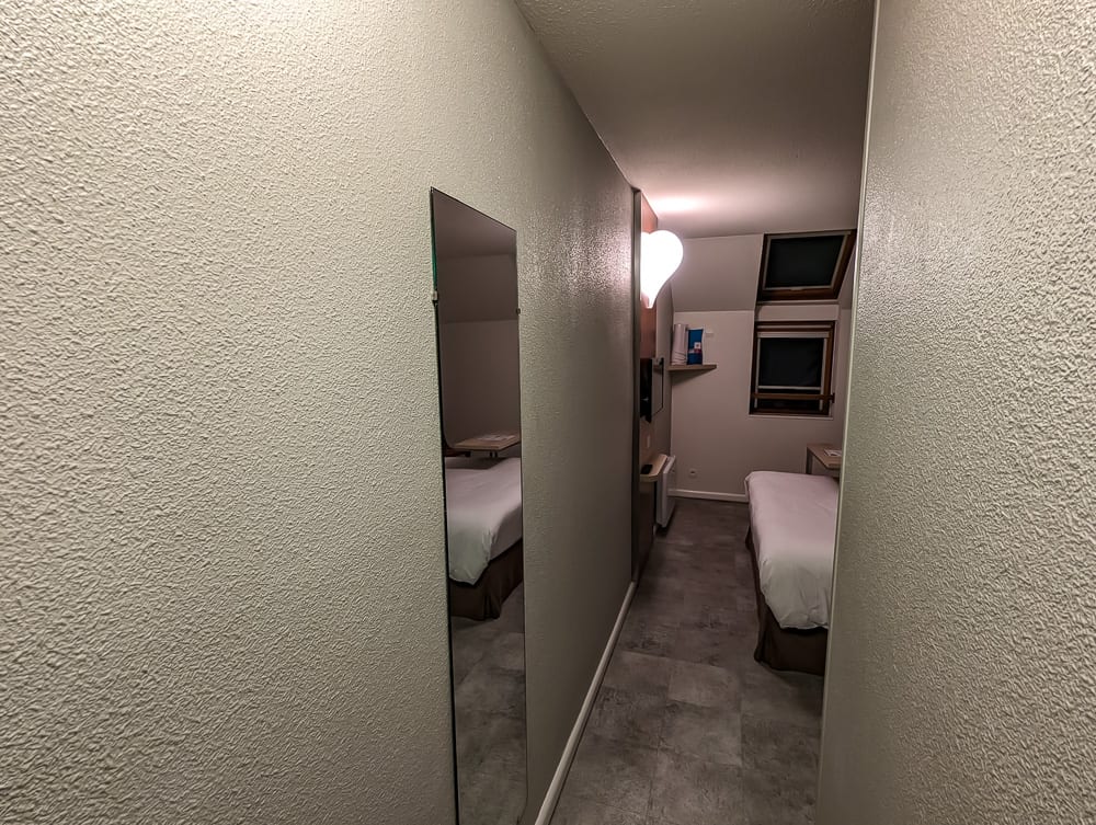 Couloir étroit menant à une petite chambre avec lit simple, miroir mural et deux fenêtres de toit