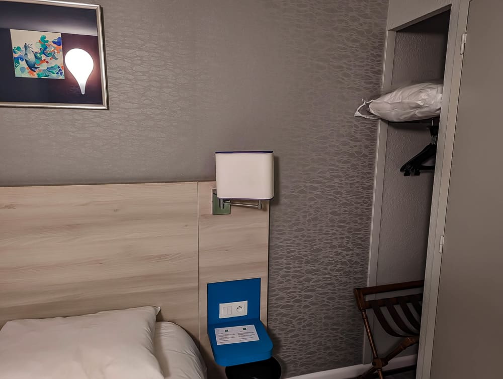 Tête de lit en bois clair avec lampe murale, petit coin penderie et coussin supplémentaire à l'Ibis Styles