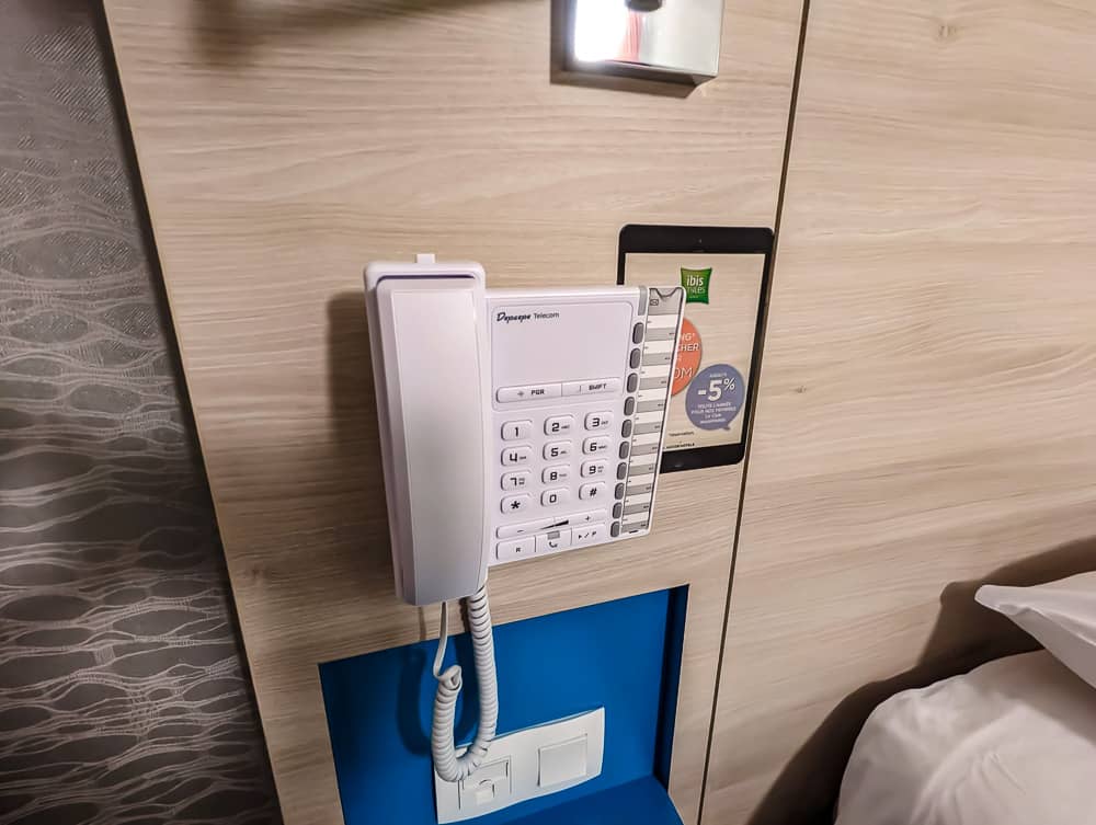 Téléphone blanc fixé au mur en bois clair près du lit d'une chambre Ibis Styles Ouistreham