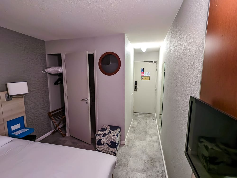 Couloir étroit d'une chambre Ibis Styles Ouistreham avec porte d'entrée, penderie ouverte et miroir mural