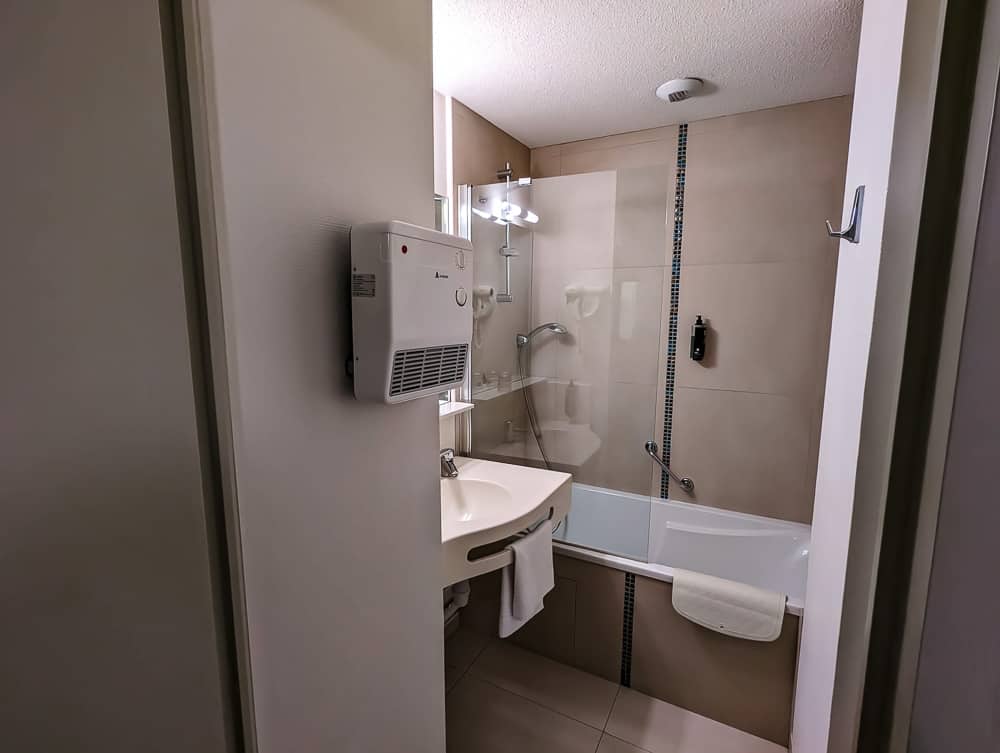 Salle de bain compacte avec baignoire, lavabo blanc, miroir éclairé et sèche-cheveux mural à l'Ibis Styles Ouistreham