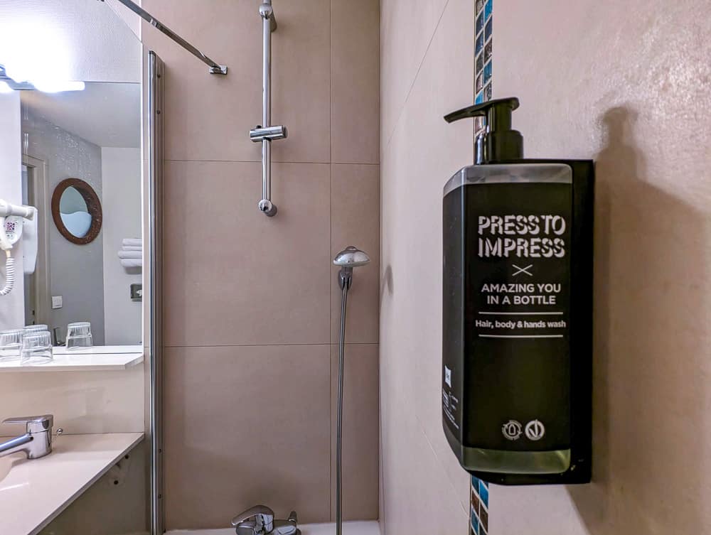 Distributeur de savon noir fixé au mur d'une salle de bain d'hôtel avec douche et miroir à l'Ibis Styles