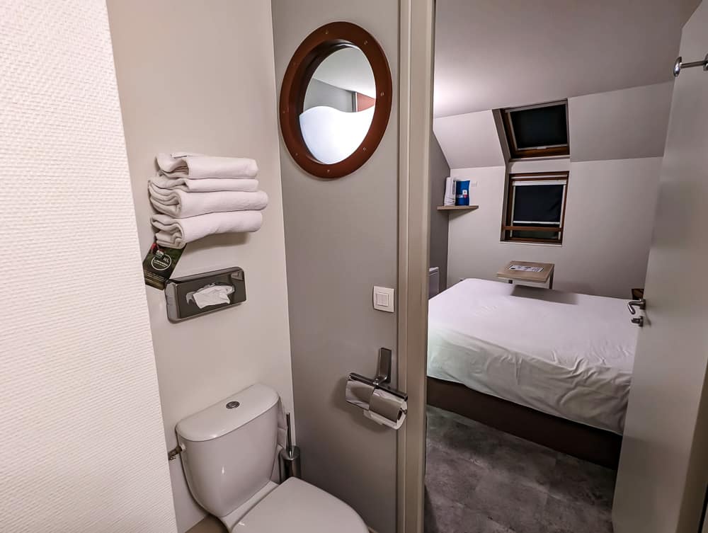Petite salle de bain avec toilettes et serviettes donnant sur une chambre mansardée à lit double