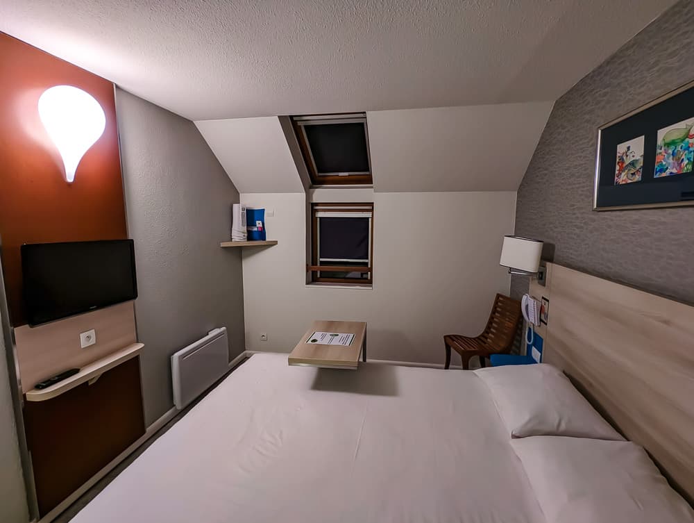 Chambre compacte à lit double avec lucarne, bureau mural, télévision et chaise en bois à l'Ibis Styles Ouistreham