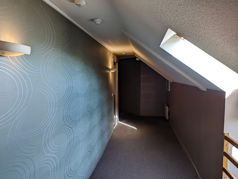 Couloir mansardé de l'hôtel Ibis Styles Ouistreham avec papier peint à motifs circulaires et lumière naturelle