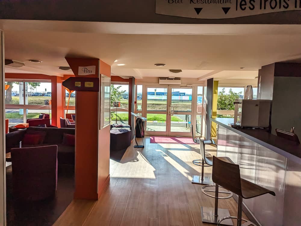 Espace bar lumineux de l'Ibis Styles Ouistreham avec comptoir blanc, chaises hautes et fauteuils violets