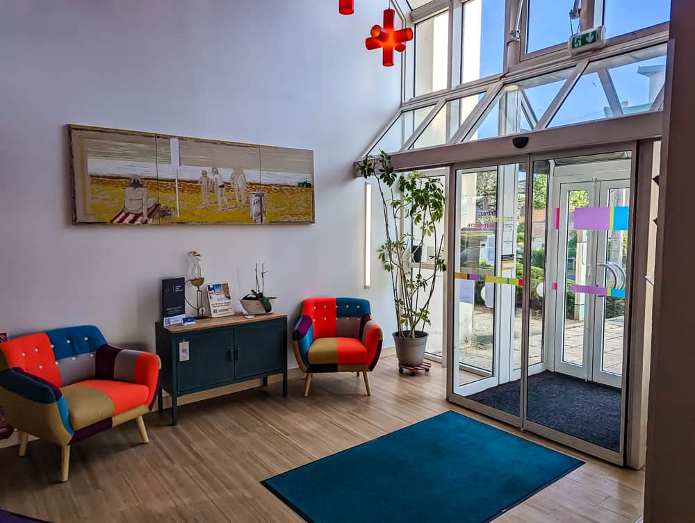 Hall d'entrée lumineux avec deux fauteuils colorés, tableau de plage et grandes portes vitrées à l'Ibis Styles