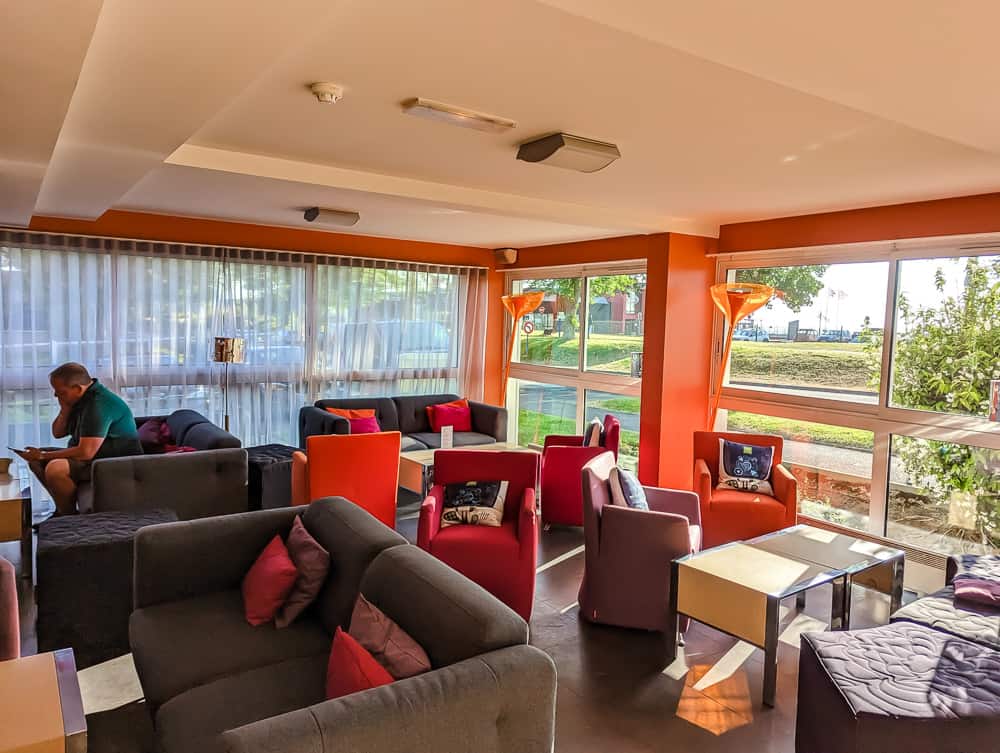 Salon lumineux de l'hôtel Ibis Styles Ouistreham avec fauteuils colorés, grandes baies vitrées et client lisant