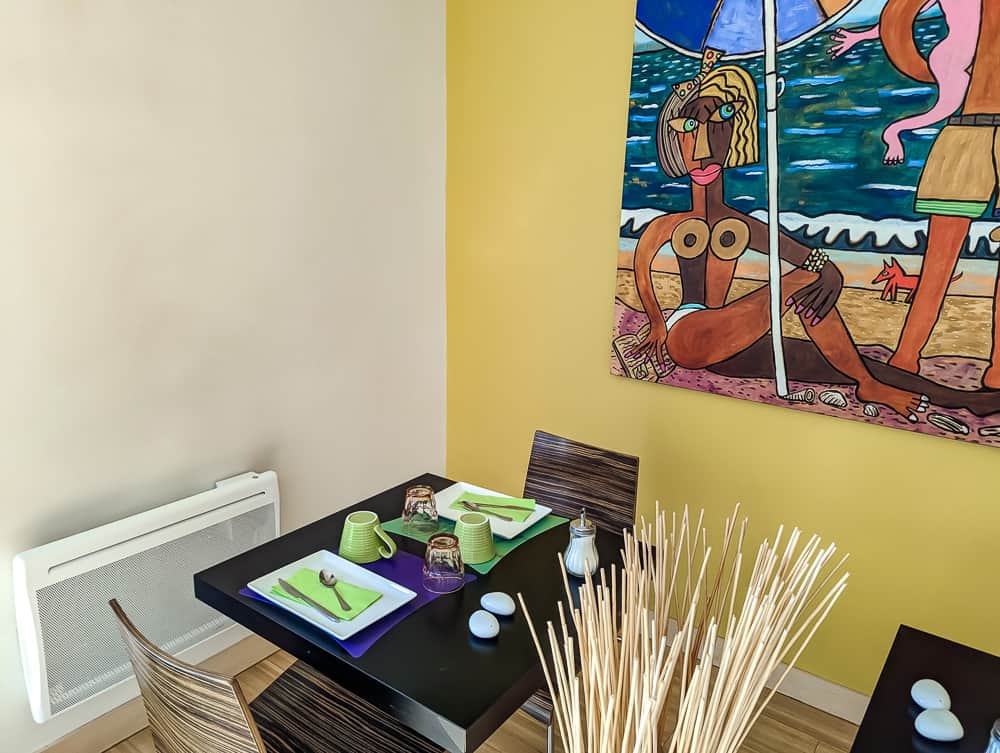 Coin repas coloré avec table noire, vaisselle verte et grand tableau balnéaire sur mur jaune à l'Ibis Styles