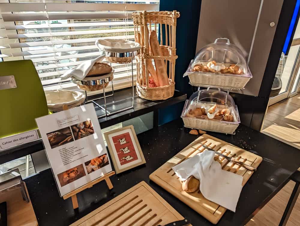 Buffet petit-déjeuner de l'Ibis Styles Ouistreham avec pains, croissants et présentoirs en osier près d'une fenêtre
