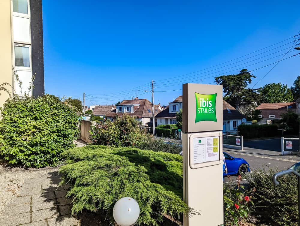 Panneau d'entrée de l'hôtel Ibis Styles Ouistreham entouré de verdure et de maisons de quartier