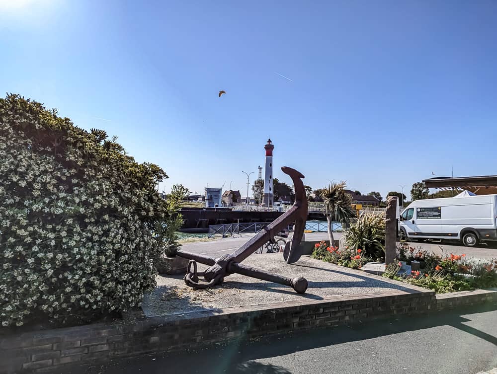 Grande ancre en métal rouillé devant un phare rouge et blanc sous un ciel bleu clair à Ouistreham