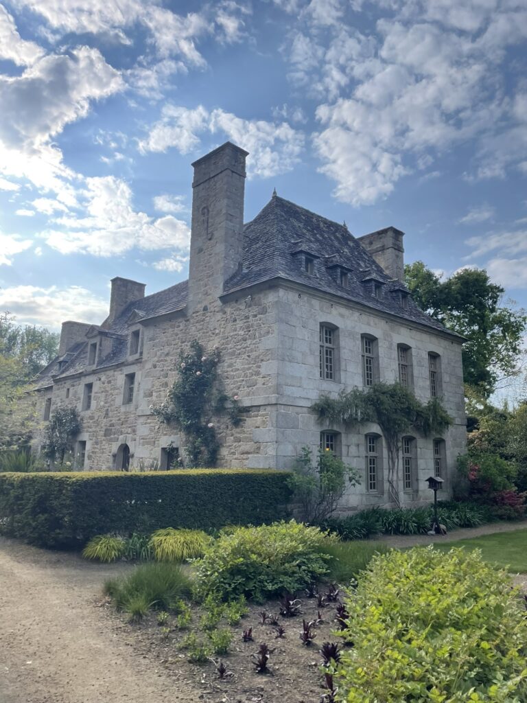 Maison en pierre grise entourée de verdure et de massifs fleuris sous un ciel partiellement nuageux.
