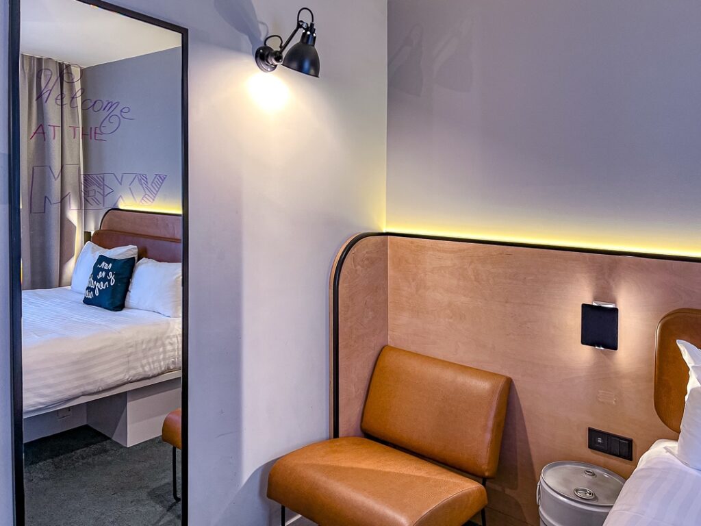 Chambre moderne du Moxy Paris Bastille avec miroir, fauteuil en cuir caramel et lit reflété.
