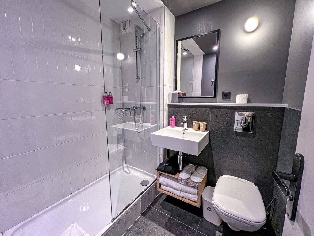 Salle de bain moderne du Moxy Paris Bastille avec douche vitrée, murs clairs, lavabo suspendu et WC.
