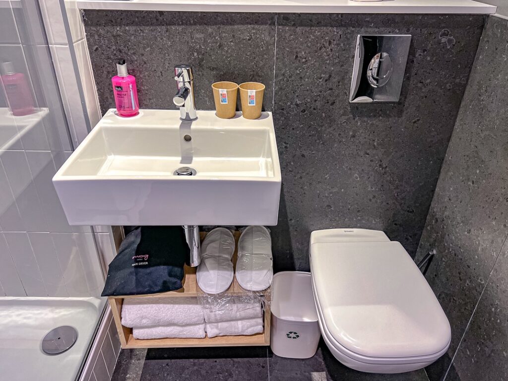 Petite salle de bain moderne du Moxy Paris Bastille avec lavabo blanc, toilettes et accessoires soigneusement rangés.