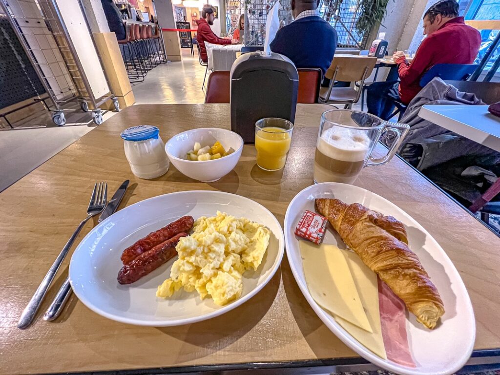 Petit-déjeuner complet au Moxy Paris Bastille avec œufs brouillés, saucisses, croissant, jambon, fromage et café latte.
