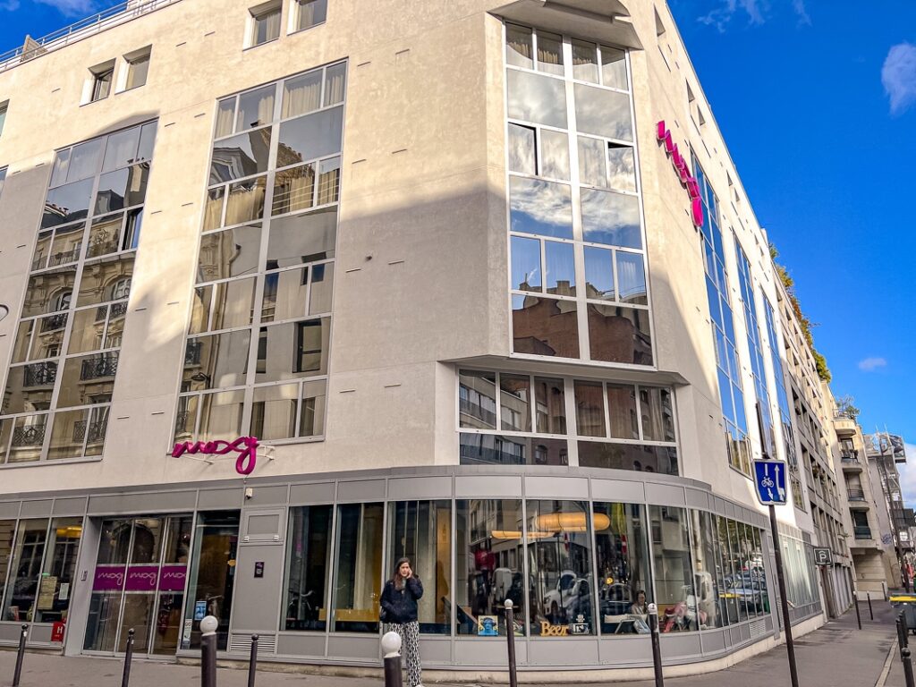 Façade moderne du Moxy Paris Bastille avec larges vitres reflétant les immeubles voisins et ciel bleu.