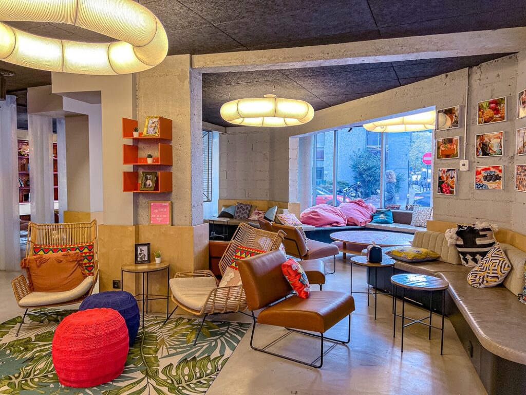 Salon coloré du Moxy Paris Bastille avec fauteuils en rotin, coussins vifs et grandes lampes circulaires suspendues.