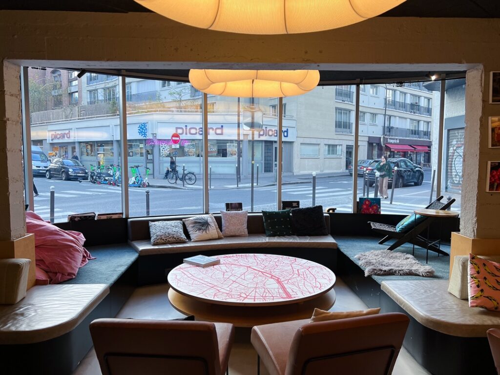 Salon convivial du Moxy Paris Bastille avec grandes baies vitrées, table ronde design et coussins colorés.