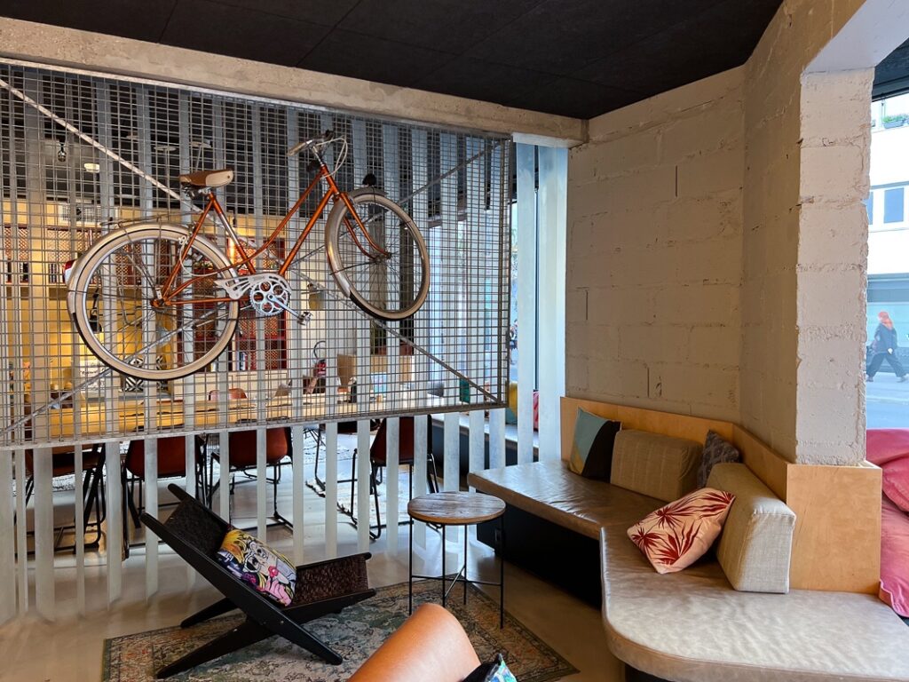 Salon moderne du Moxy Paris Bastille avec vélo suspendu, canapés beiges et chaise design colorée.